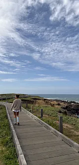 A woman walks along a wooden boardwalk toward the rocky ocean under dramatic cloudy skies.