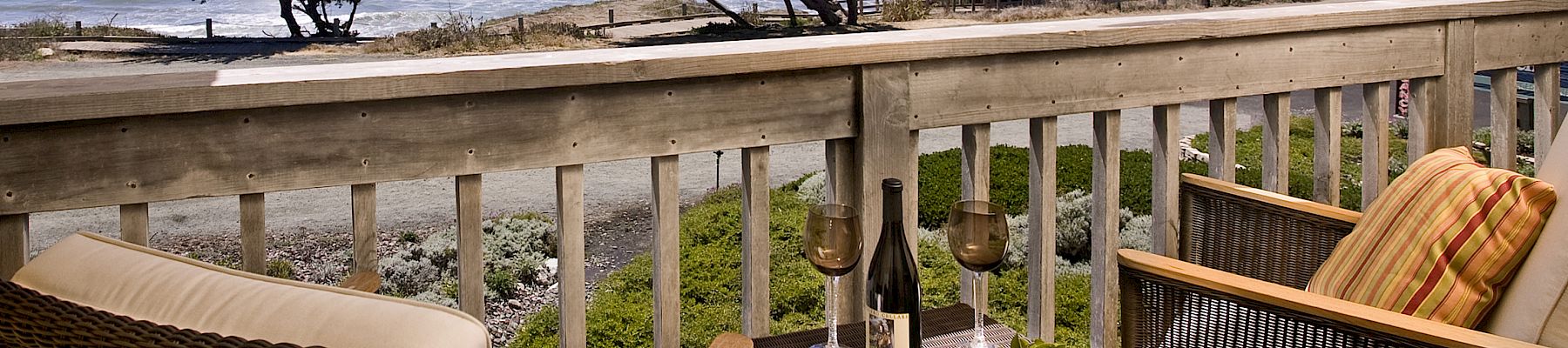 Balcony dining setup with wine, chairs, and a view of the ocean, rocky shore, waves, and coastal trees under a blue sky.