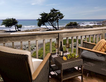 Balcony dining setup with wine, chairs, and a view of the ocean, rocky shore, waves, and coastal trees under a blue sky.