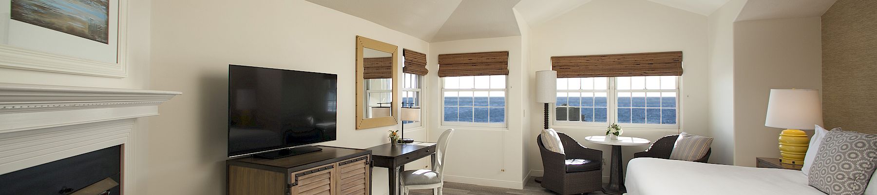 A bright hotel bedroom with ocean-view windows, blinds, a TV on a dresser, a fireplace, and a neatly made bed.