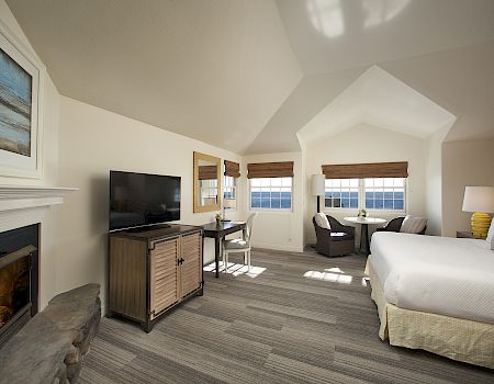 A bright hotel bedroom with ocean-view windows, blinds, a TV on a dresser, a fireplace, and a neatly made bed.