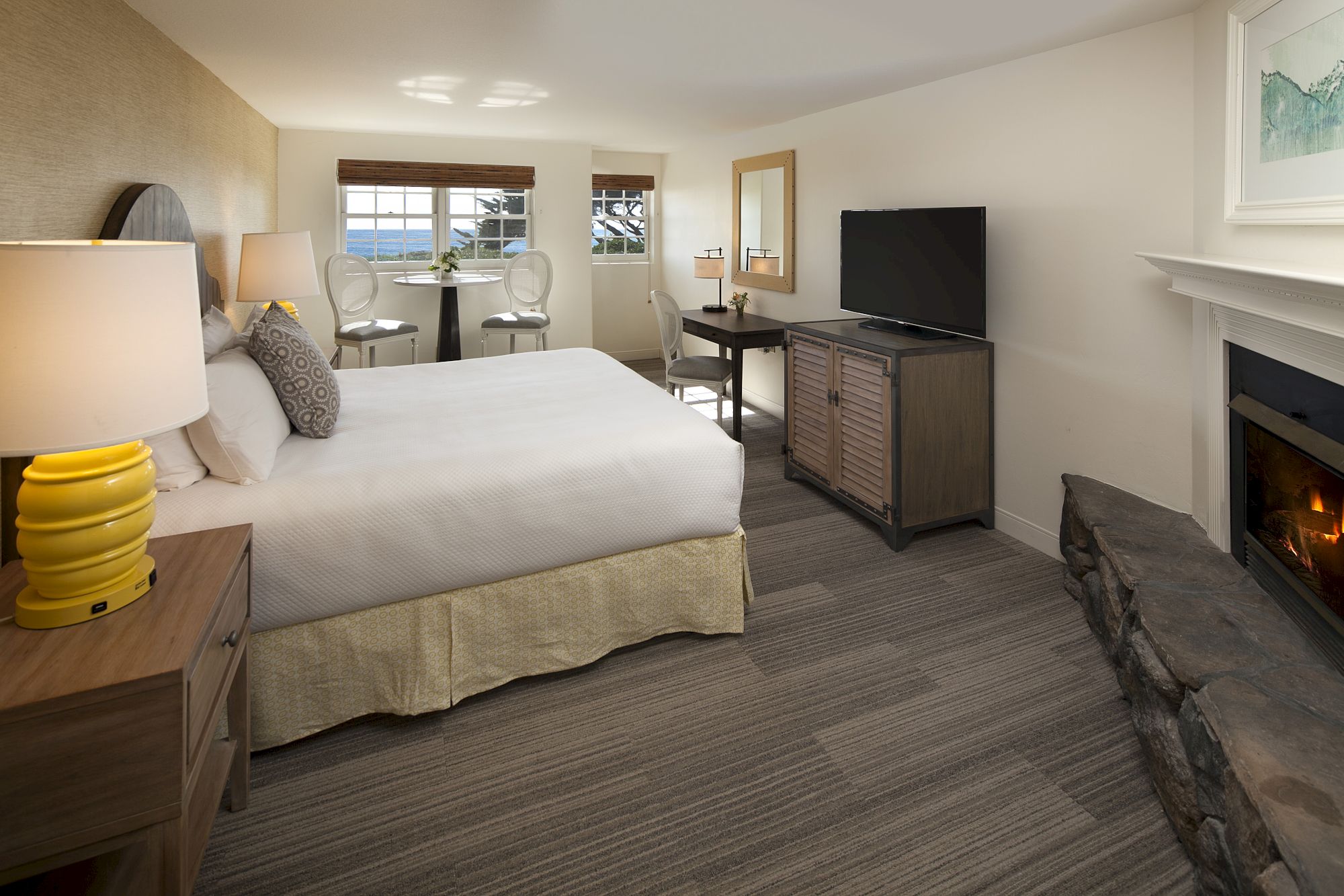 A cozy hotel room with a king bed, nightstands, desk, fireplace, TV on a dresser, and a seating area by large windows; warm, neutral tones.