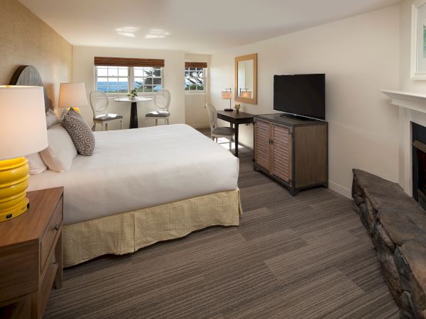 A cozy hotel room with a king bed, dresser, TV, fireplace, desk, and a small seating area by the window, all in neutral tones.