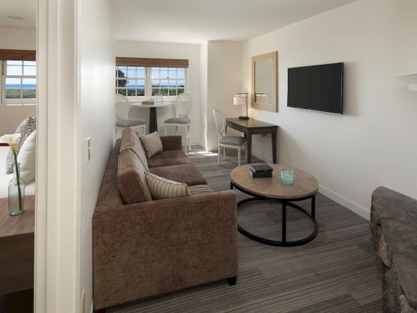 A cozy living room with a brown sofa, round coffee table, fireplace, wall TV, and a doorway opening to a bright bedroom area with windows. Top it at 140 characters, always ending the sentence.