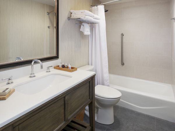 A modern bathroom with a double-sink vanity, large mirror, soap and towels, a toilet, and a bathtub with a shower curtain and grab bar, all in neutral tones.