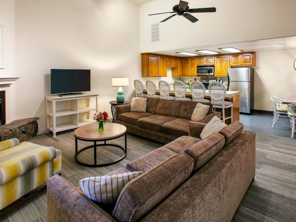 Spacious open-concept living room with a brown sectional, patterned chair, coffee table, fireplace, TV stand, and a bright kitchen/dining area in the background.