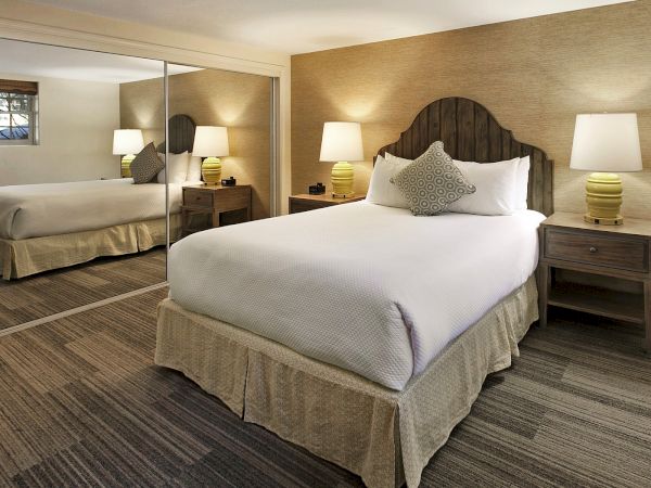 A tidy hotel room with a large bed, mirrored closet, nightstands, lamps, and a neutral beige decor, all under warm lighting.