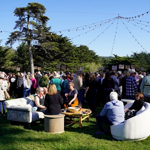 People gathered at an outdoor party or festival on a sunny day, mingling around blankets and seating with string lights overhead.