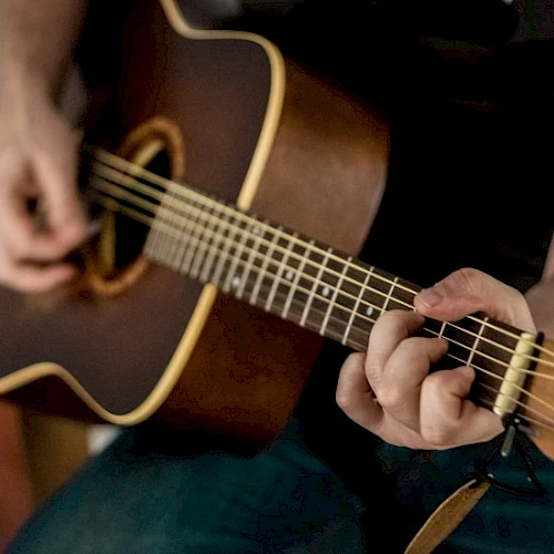 A person playing a brown acoustic guitar, fingers strumming on the strings while the other hand frets near the neck.