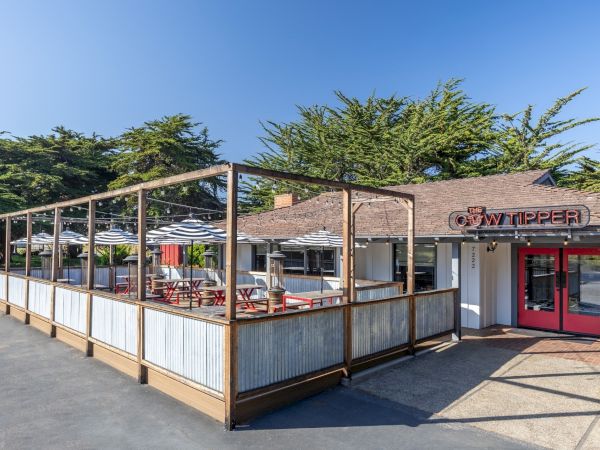A small outdoor dining area attached to a rustic wooden-framed restaurant with red doors, patio tables, and a bright, sunny day.