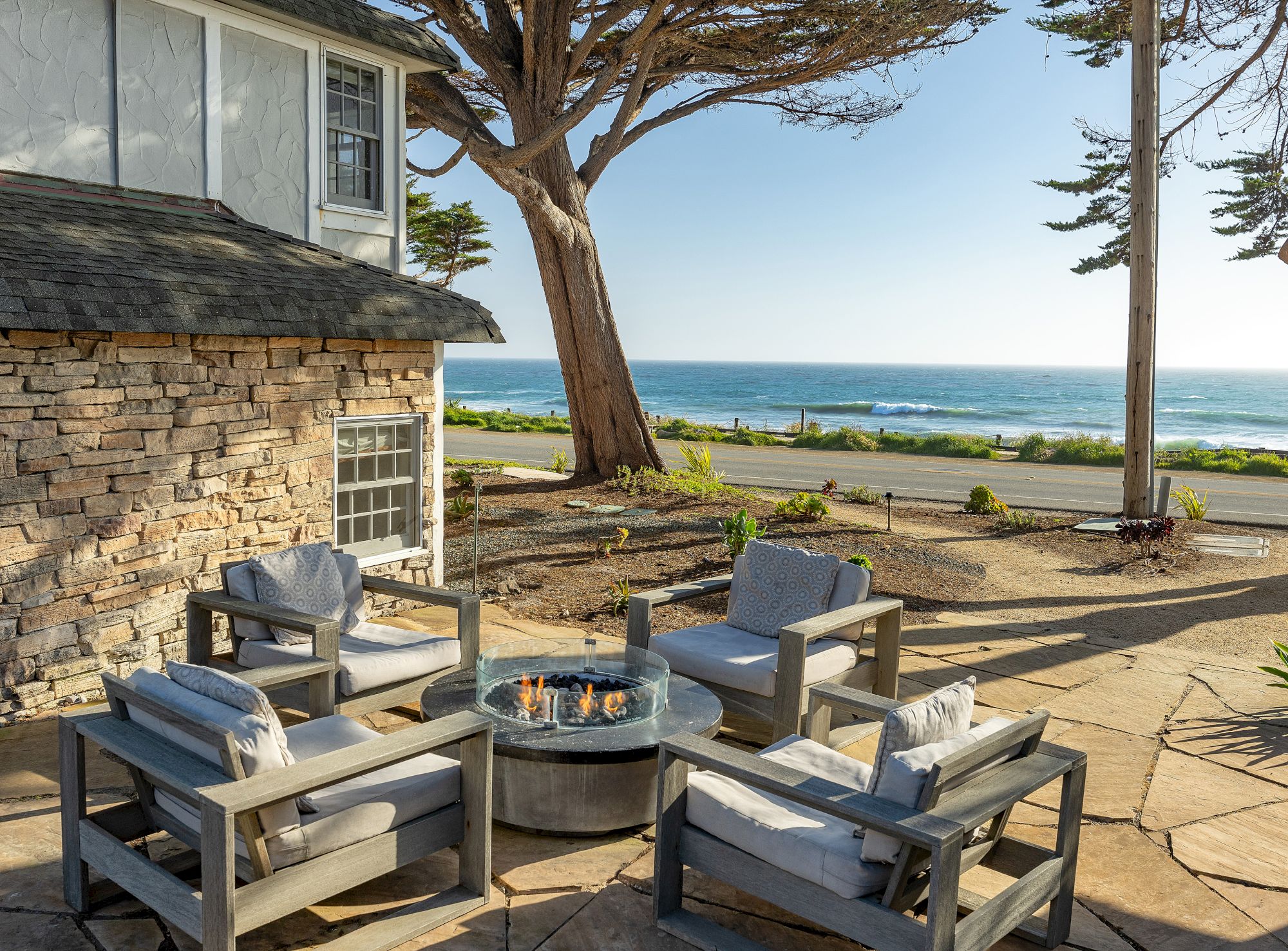 A seaside patio with comfy gray chairs around a fire pit, overlooking the ocean and palm trees, near a stone house by the shore.