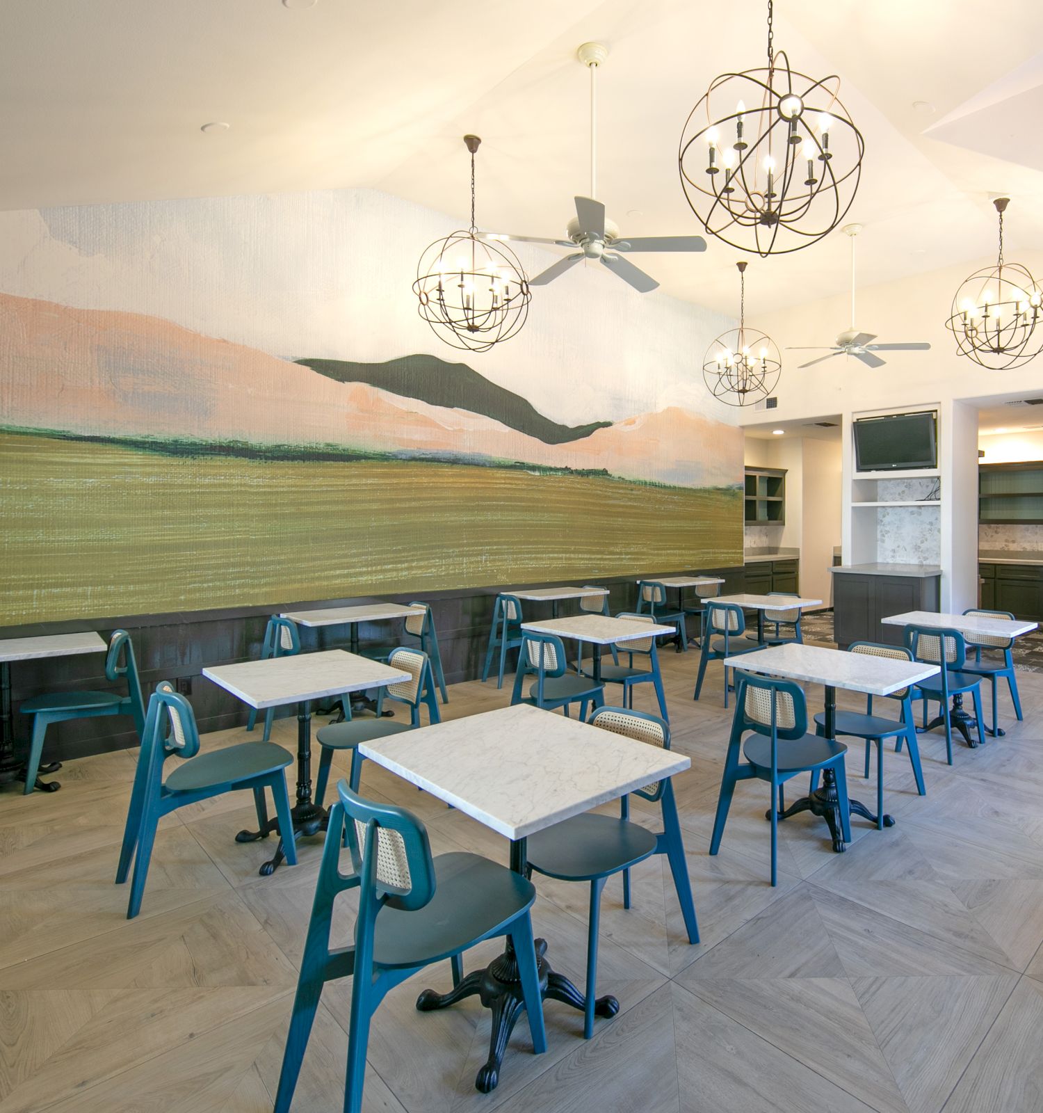 Bright, modern cafe with blue metal chairs, white marble tables, a long mural of a beach and hills on the left, and decorative spherical light fixtures.