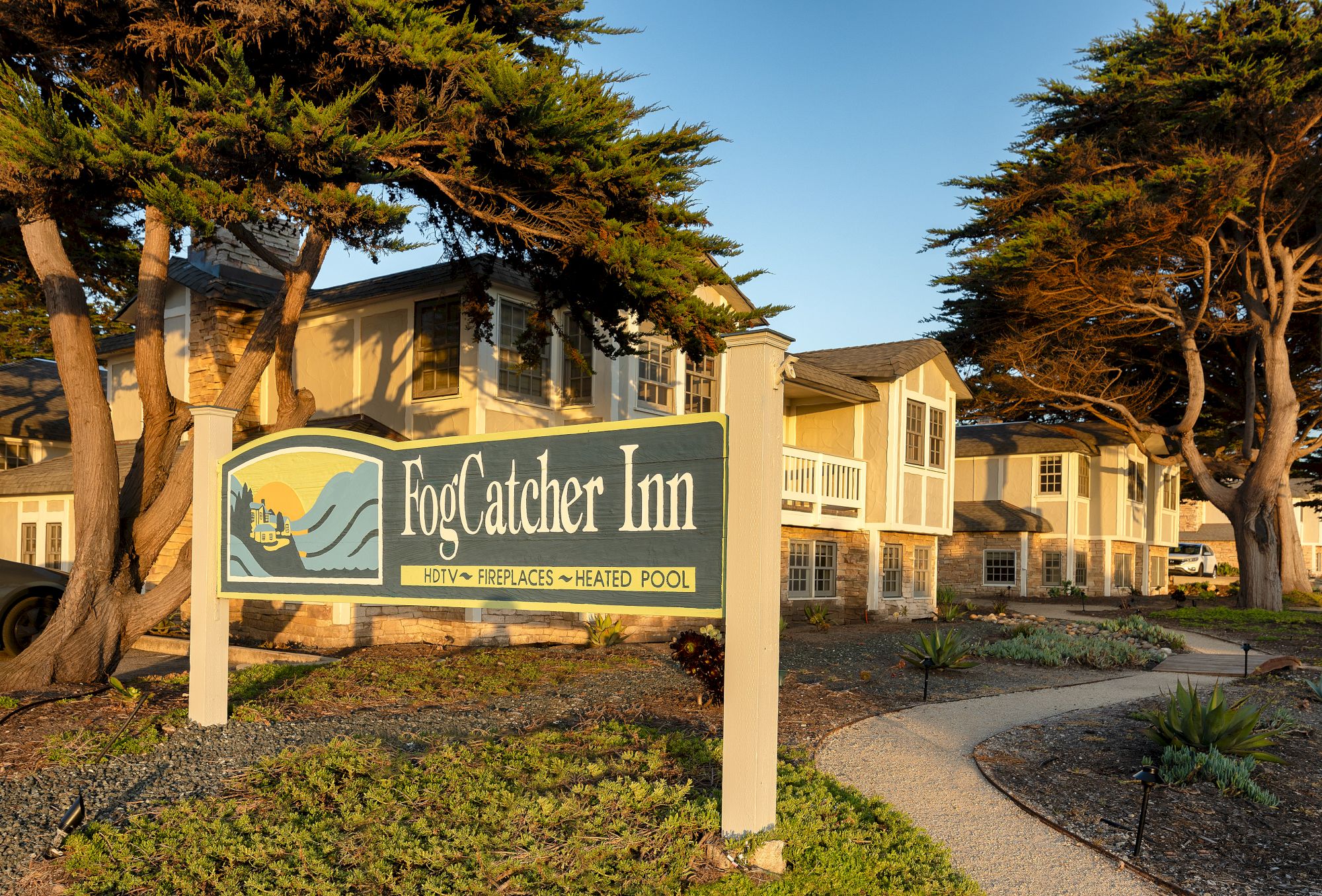 A FogCatcher Inn sign outside a coastal property with path, trees, and beige buildings in the background.