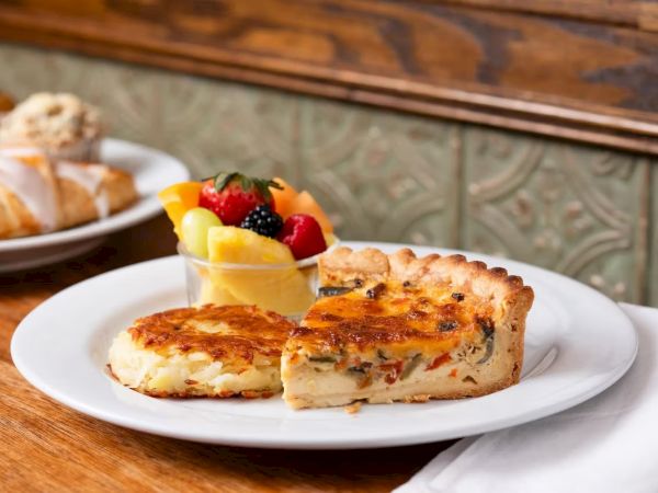 A plated slice of quiche with a golden crust, a smaller portion, and a side of fresh fruit parfait on a white plate.