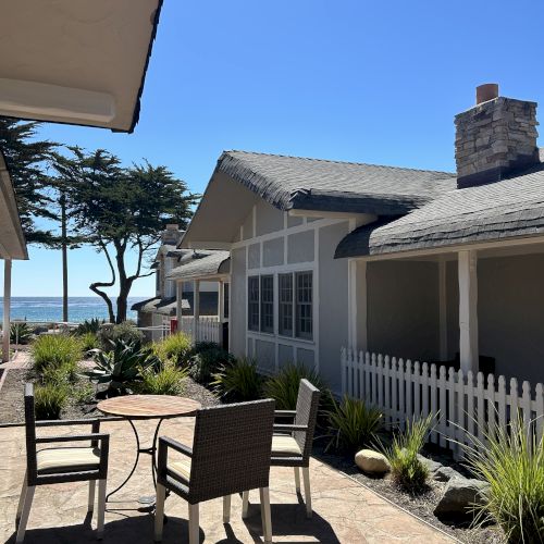 A sunny beachfront patio with wicker seating, a white picket fence, tropical plants, and cozy cottages by the ocean under a clear blue sky.
