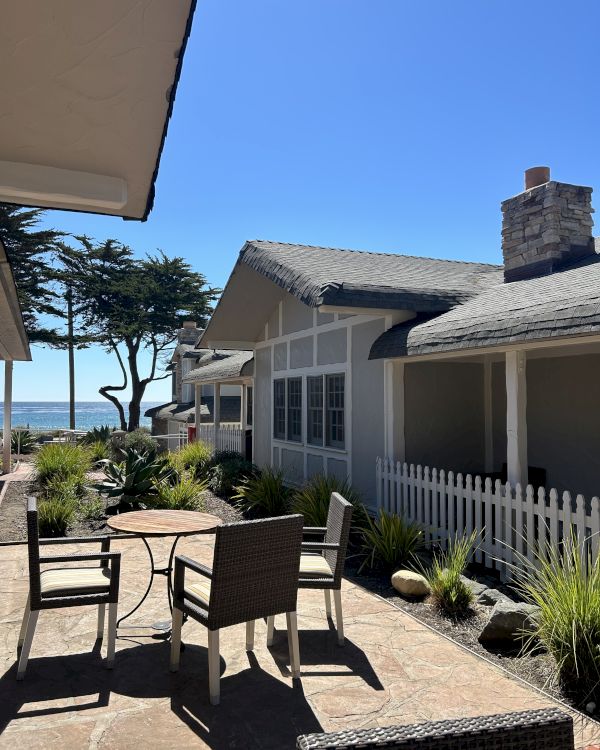 A sunny beachfront patio with wicker seating, a white picket fence, tropical plants, and cozy cottages by the ocean under a clear blue sky.