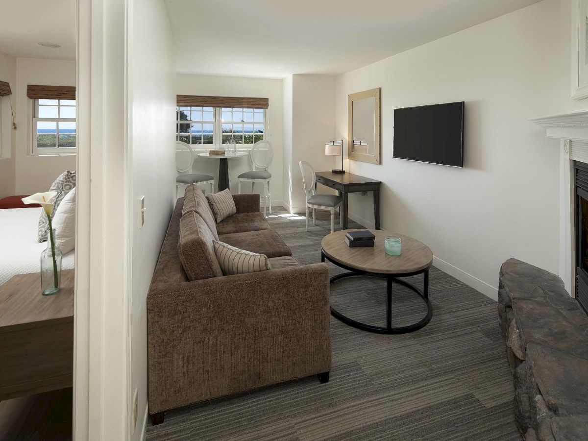 A modern hotel suite with a cozy living area: brown sofa, round coffee table, wall-mounted TV, fireplace, and a bedroom glimpsed through an open doorway.