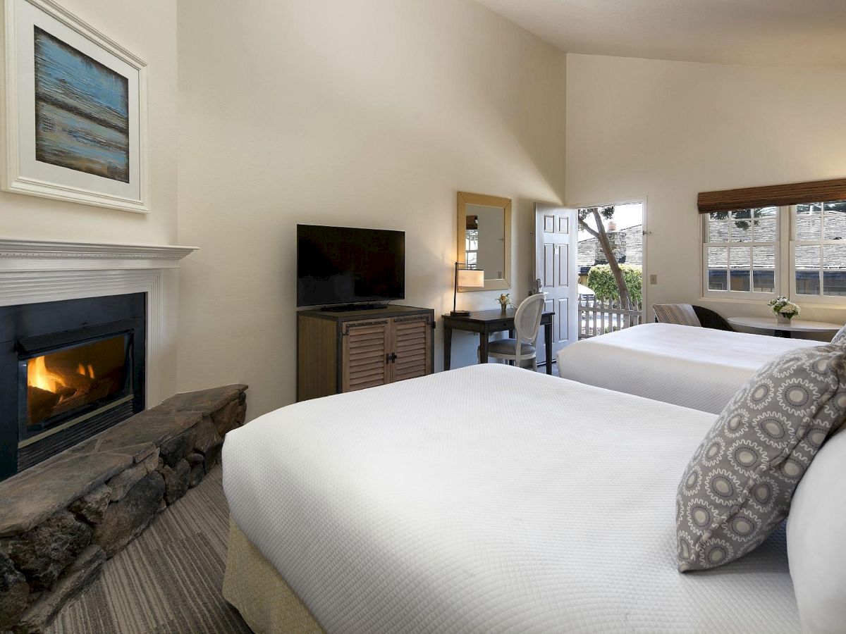 A cozy hotel room with two beds, a fireplace, desk, TV, and a small seating area by a window, creating a warm, inviting stay.