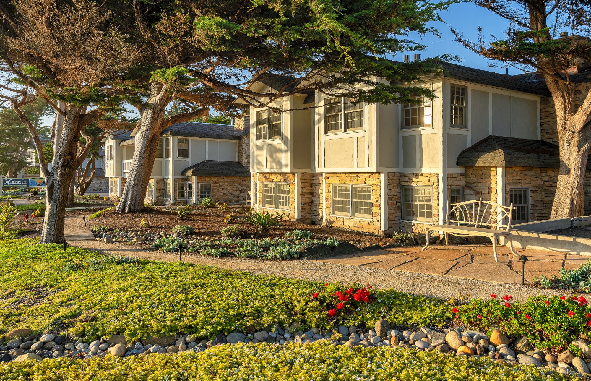 A row of beige two-story townhouses with stone bases, large trees, a manicured front yard, and a sunny, tranquil residential street.