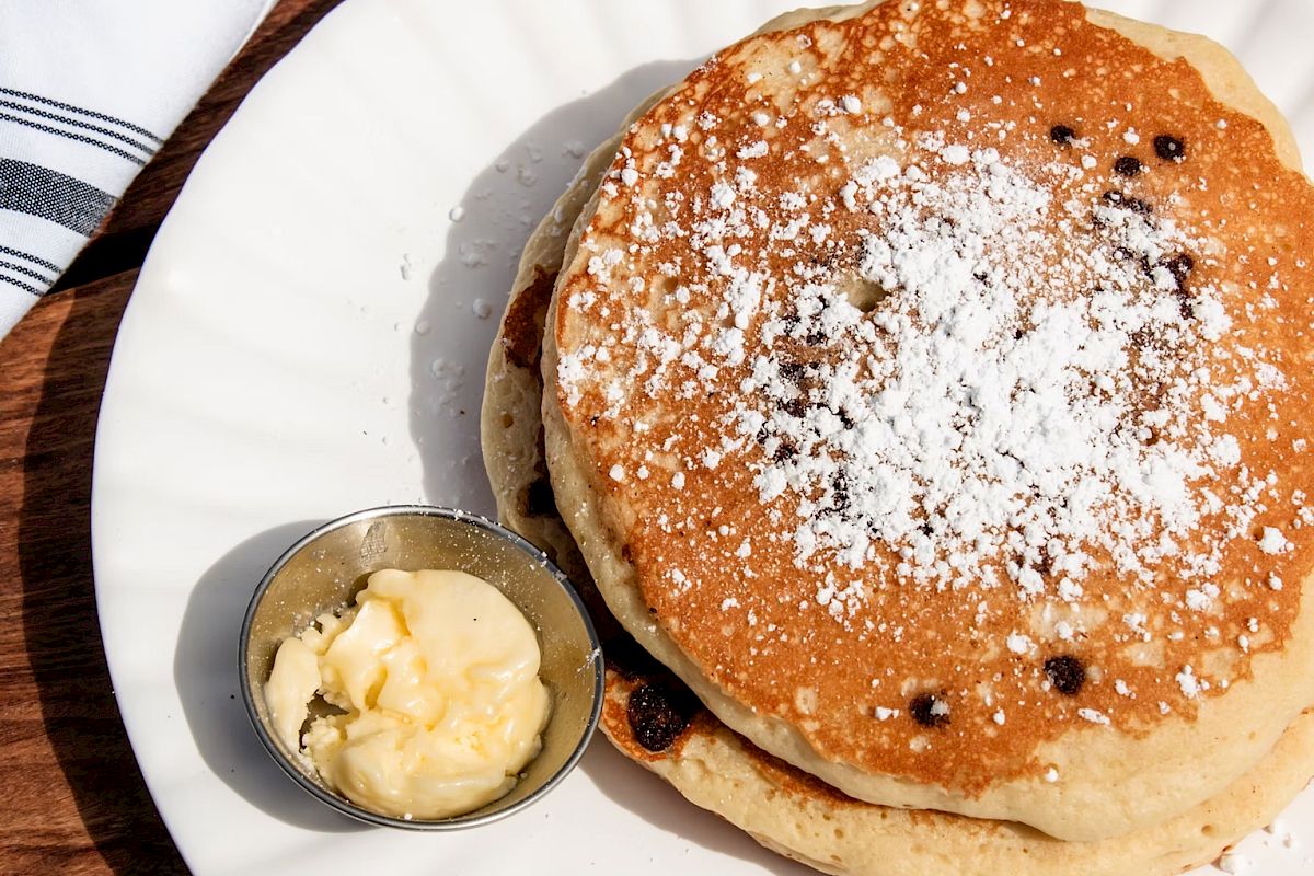 A stack of fluffy pancakes with powdered sugar on a white plate, served with a small cup of butter. Delicious breakfast treat.