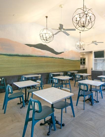Bright cafe with mint chairs, square tables, mural of hills, modern light fixtures, and a calm, airy layout.