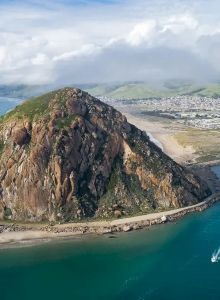 Aerial view of a coastal landscape with a prominent rocky hill rising from the sea, surrounded by turquoise water and a sandy shore.