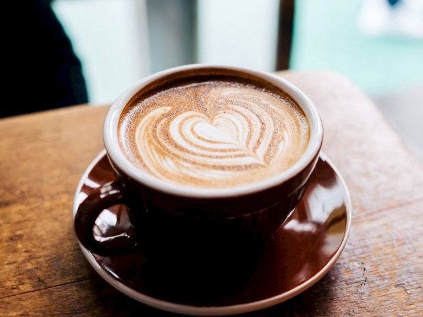 A brown ceramic cup of latte on a wooden table, topped with a heart-shaped foam latte art design, with a saucer underneath.
