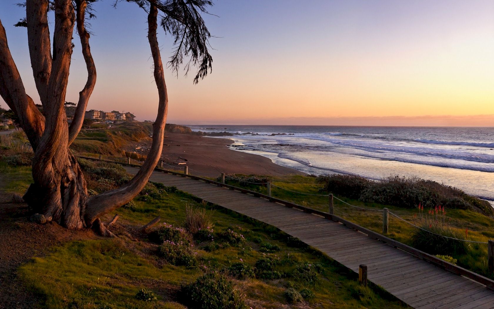 A coastal sunset scene with a wooden path along the beach, trees on the left, calm waves, and warm orange-pink sky ending at the horizon.