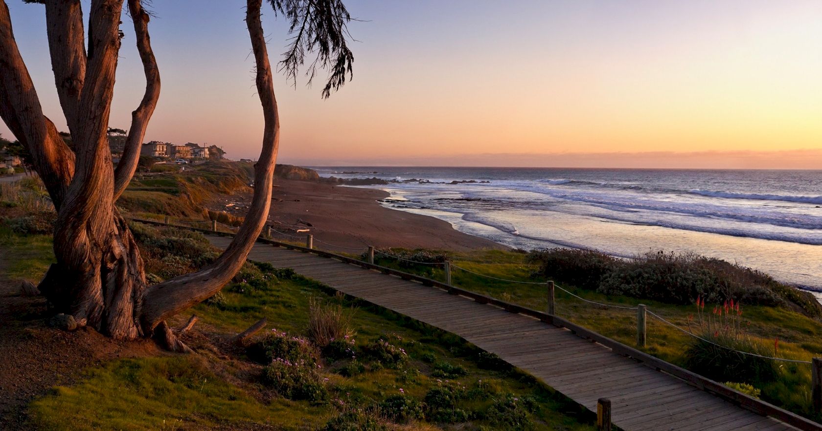 A coastal sunset scene with a wooden path along the beach, trees on the left, calm waves, and warm orange-pink sky ending at the horizon.
