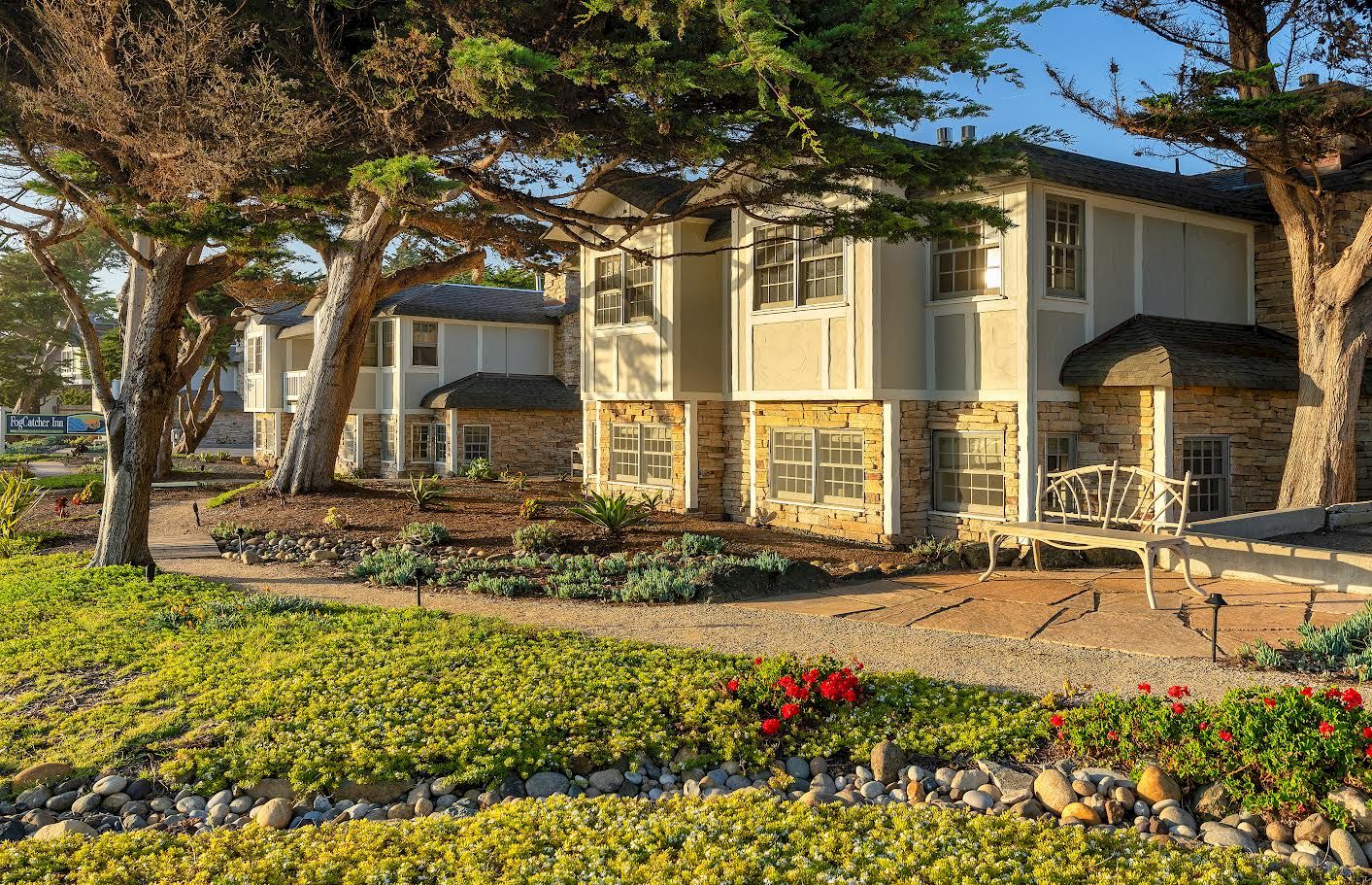 A tidy townhouse complex with beige multi-story buildings, stone accents, manicured lawns, trees lining the walkway, and a sunny, welcoming atmosphere.