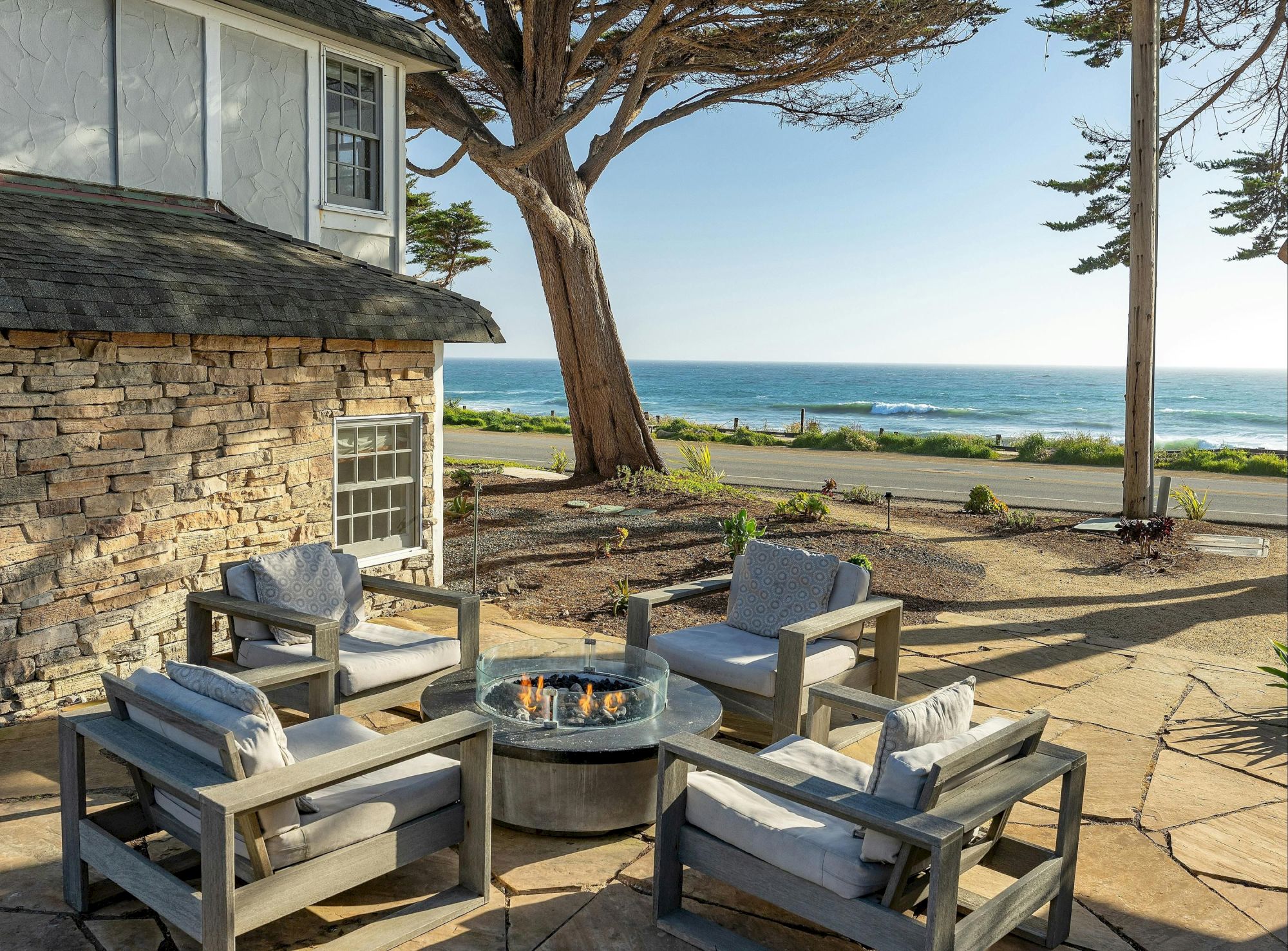 A stylish outdoor seating area with cushioned chairs around a fire pit on a stone patio, overlooking a sandy beach and ocean beyond.