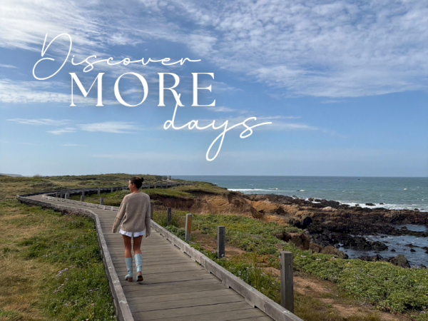 A person walks a wooden boardwalk along a rugged ocean coastline under cloudy skies, with &ldquo;Discover more days&rdquo; text overlaid.