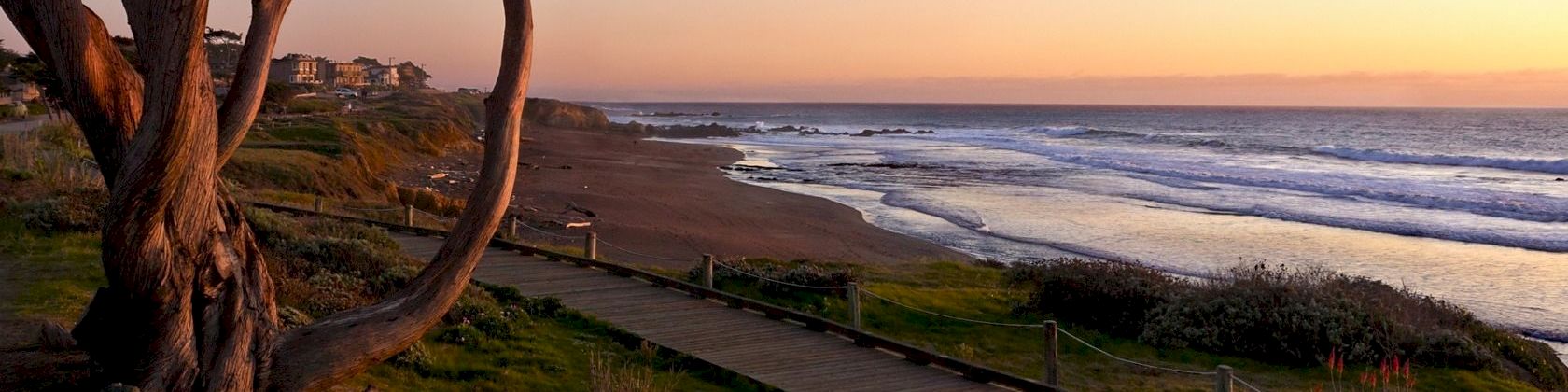 A sunset beach with crashing waves, a wooden boardwalk along the sand, and tall trees framing the shoreline.