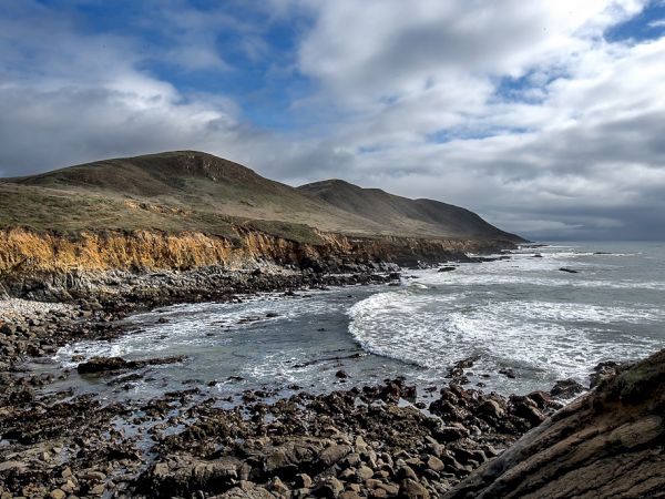 Coastal cliffs meet a rocky shore as waves crash along the rugged shoreline under a partly cloudy sky.