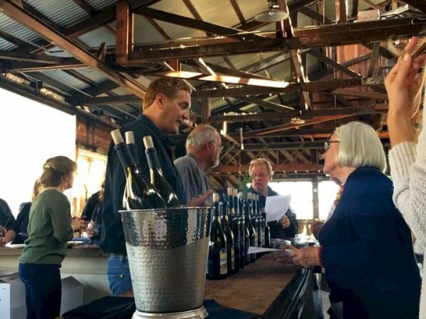 People at a rustic bar tasting bottles; a man pours, others chat and sample drinks under wooden beams.