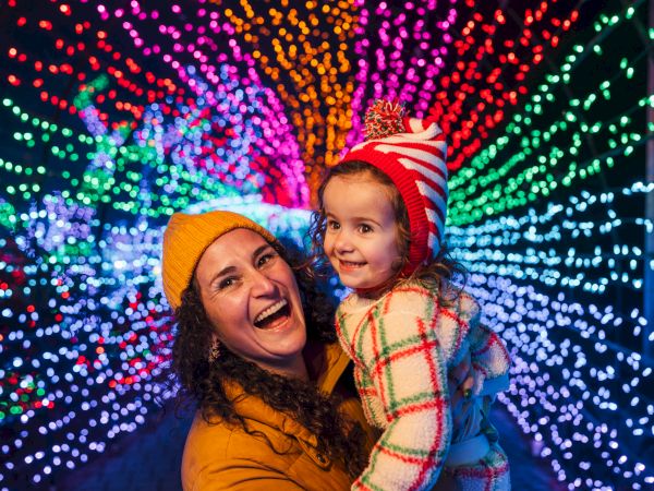 A joyful mom holds a smiling little girl in winter hats beneath colorful festival lights and bokeh sparkles around them.