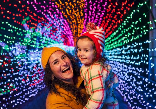 A woman holds a smiling child in winter hats as colorful holiday lights sparkle behind them.