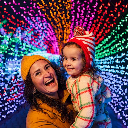 A woman holds a smiling child in winter hats as colorful holiday lights sparkle behind them.