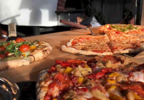 A pizzaiolo slides colorful pizzas from a wood-fired oven onto a peel, with other pies in the background and ready to bake.
