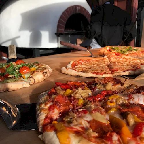 A pizzaiolo slides colorful pizzas from a wood-fired oven onto a peel, with other pies in the background and ready to bake.