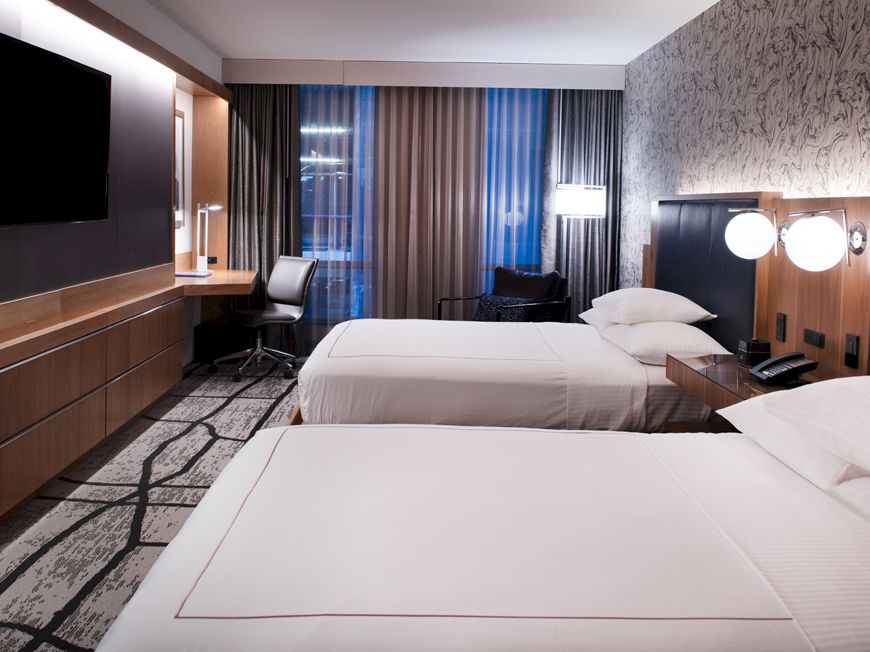 A modern hotel room with two beds, wood paneling, curtains, and wall-mounted TVs.
