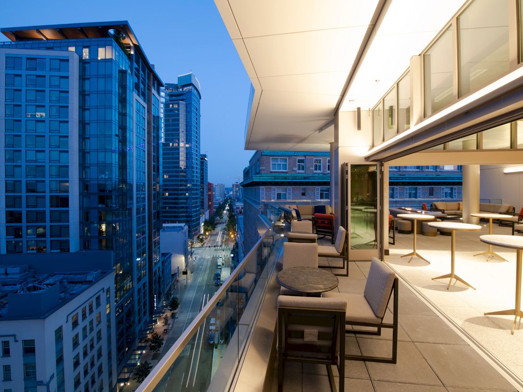 Modern high-rise rooftop terrace overlooks a city street with lounge seating and tall buildings at dusk.